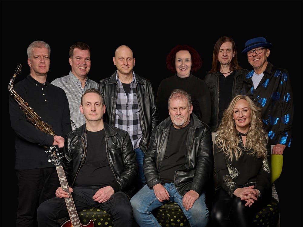 a band, with various instruments visible including a saxophone and guitar. The group consists of about 9-10 people arranged in two rows, with some wearing leather jackets and casual attire.