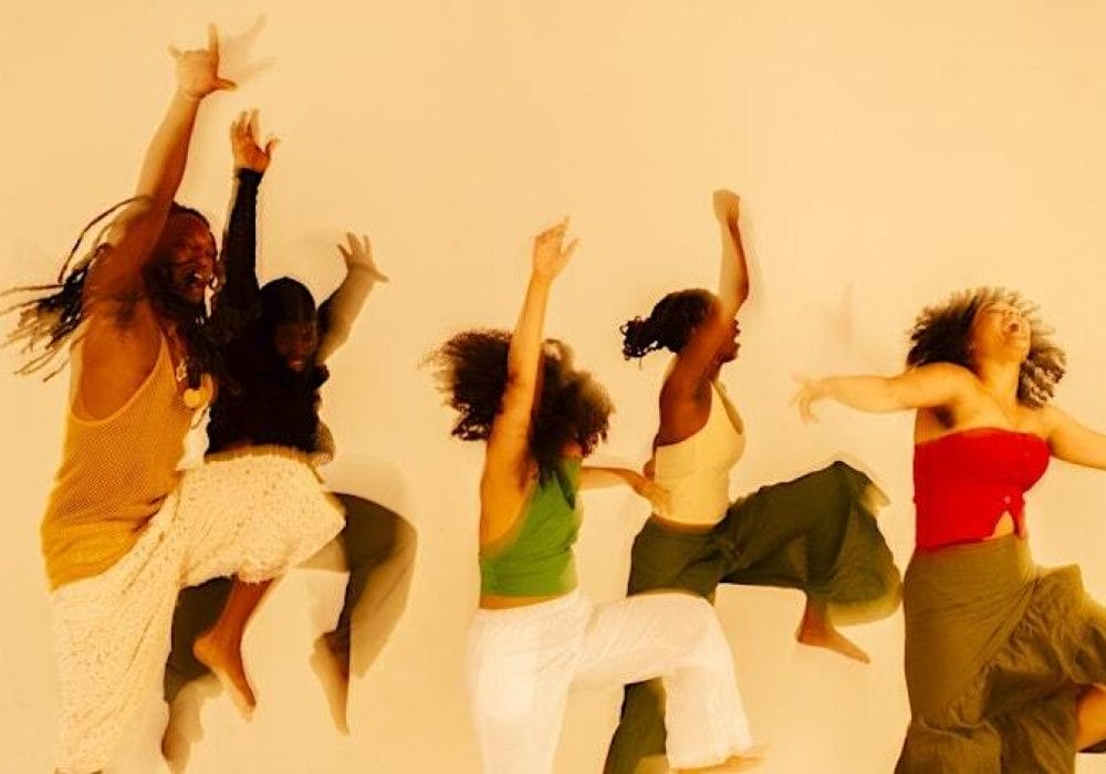 Five people dancing energetically against a plain background, dressed in vibrant and earthy-toned clothing, with expressive body movements and joyful expressions.