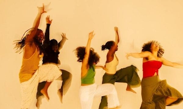 Five people dancing energetically against a plain background, dressed in vibrant and earthy-toned clothing, with expressive body movements and joyful expressions.