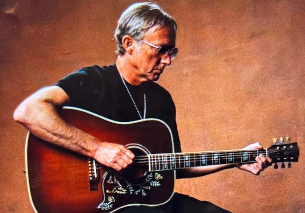A white male with short grey hair wearing a black top & trousers sat on a chair playing guitar.