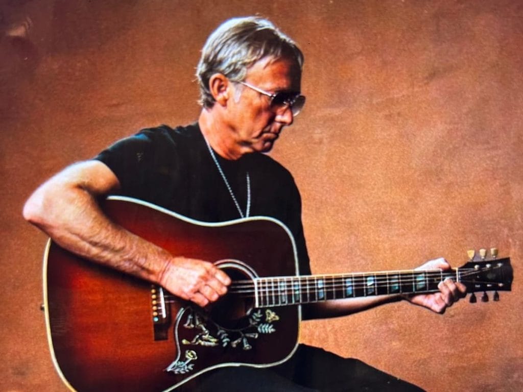 A white male with short grey hair wearing a black top & trousers sat on a chair playing guitar.