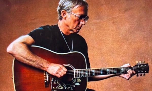 A white male with short grey hair wearing a black top & trousers sat on a chair playing guitar.