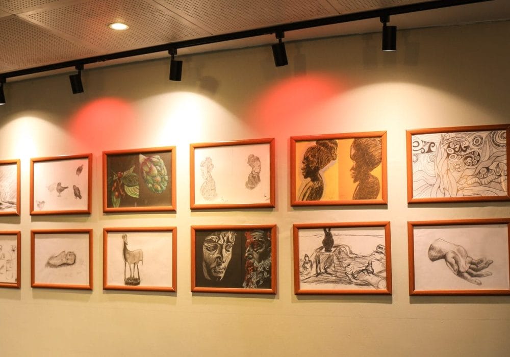 A gallery wall with twelve framed artworks arranged in two rows, each in a light brown frame. The pieces include pencil sketches, charcoal drawings, and colored illustrations, featuring subjects such as human faces, profiles, hands, animals, plants, and abstract designs. The wall is lit by ceiling spotlights, with a soft red glow over the upper middle artworks. In the background, more framed art is visible on an adjacent wall.