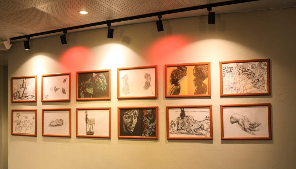 A gallery wall with twelve framed artworks arranged in two rows, each in a light brown frame. The pieces include pencil sketches, charcoal drawings, and colored illustrations, featuring subjects such as human faces, profiles, hands, animals, plants, and abstract designs. The wall is lit by ceiling spotlights, with a soft red glow over the upper middle artworks. In the background, more framed art is visible on an adjacent wall.