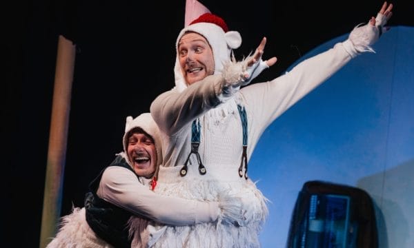 Two actors in whimsical costumes perform on a stage, one dressed in a white knit sweater and faux fur trimmed garment with suspenders, a matching hood with ear detail and a red-tipped hat, the other wearing a similar hood and a dark green jacket, both with joyful expressions. The actor in the front has one arm raised. A blue and black backdrop and a prop are partially visible.