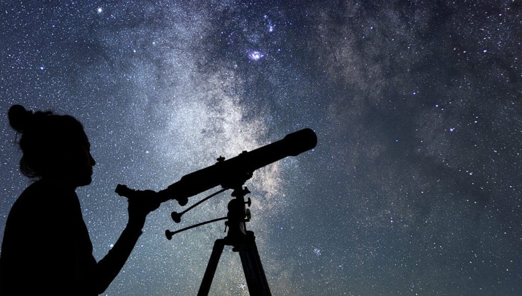 Silhouette of a person looking through a telescope at a vibrant starry night sky, Milky Way visible.