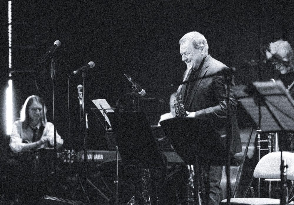 Mick laughs holding a sax while guitarist acknowledges across from him on a busy stage.