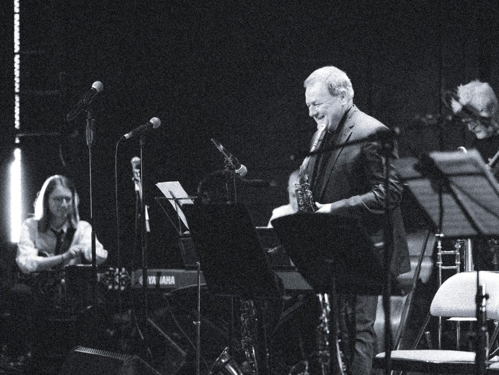 Mick laughs holding a sax while guitarist acknowledges across from him on a busy stage.