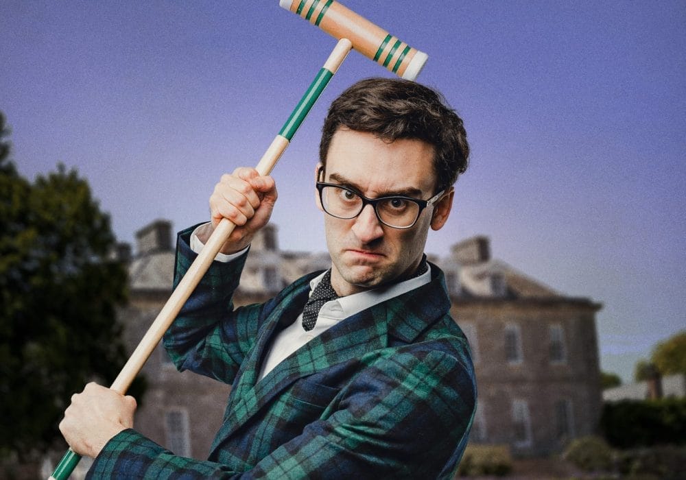 A man in formal Victorian-style attire poses with a croquet mallet raised above his shoulder. He wears black-rimmed glasses, a navy and green tartan jacket, white shirt with bow tie, and has a stern, focused expression. The background shows a blurred outdoor setting with buildings and trees under a purple-tinted sky.