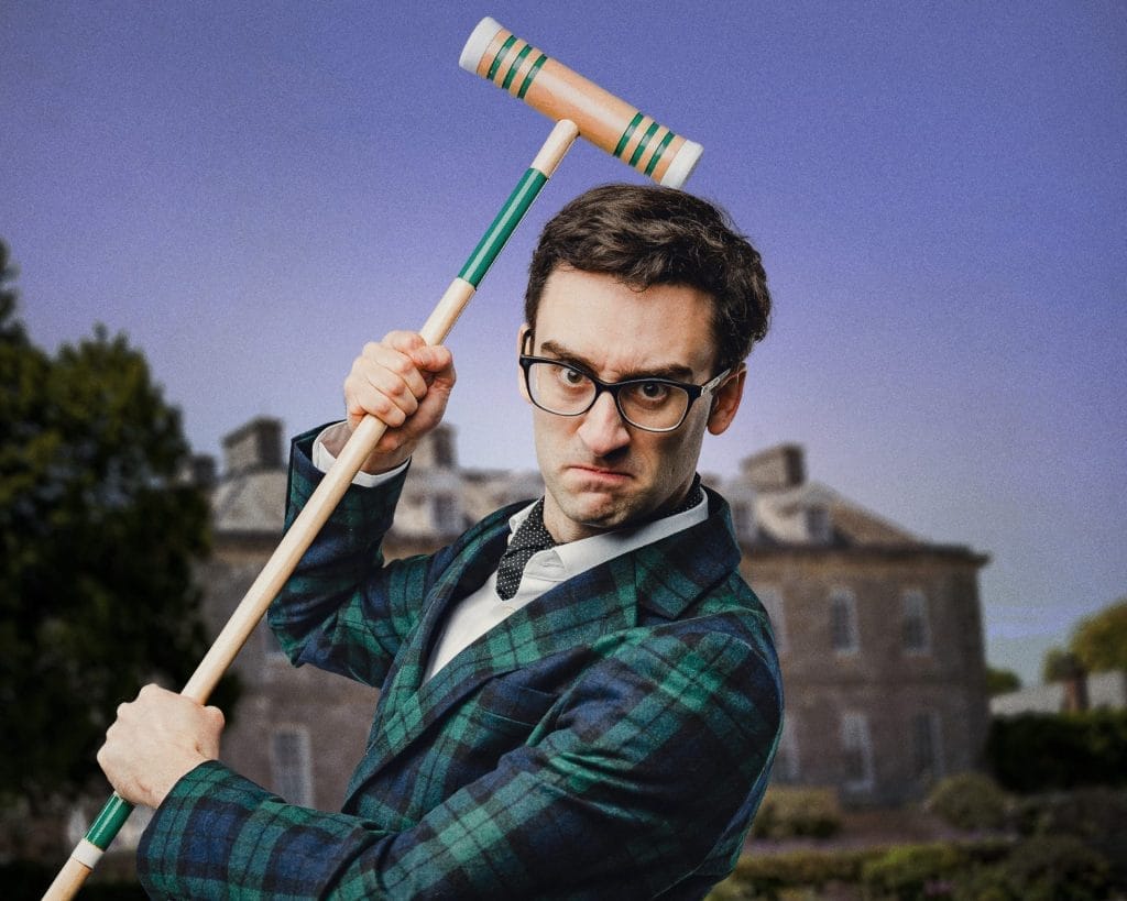 A man in formal Victorian-style attire poses with a croquet mallet raised above his shoulder. He wears black-rimmed glasses, a navy and green tartan jacket, white shirt with bow tie, and has a stern, focused expression. The background shows a blurred outdoor setting with buildings and trees under a purple-tinted sky.