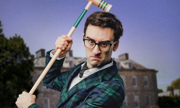 A man in formal Victorian-style attire poses with a croquet mallet raised above his shoulder. He wears black-rimmed glasses, a navy and green tartan jacket, white shirt with bow tie, and has a stern, focused expression. The background shows a blurred outdoor setting with buildings and trees under a purple-tinted sky.