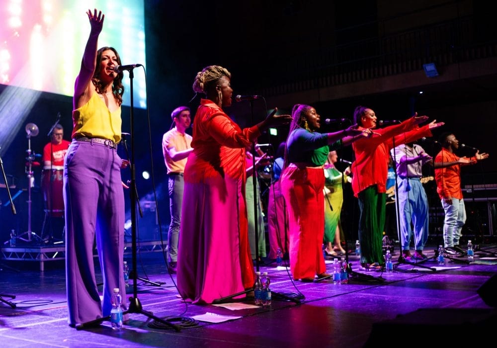 A captivating image displays a vibrant stage performance, where colorful lights and performers in electric hues (purple, pink, and red) come together, creating a spectacle of energy and joy. The images focal point is the professional bands setup, amplifying the excitement of the live show.