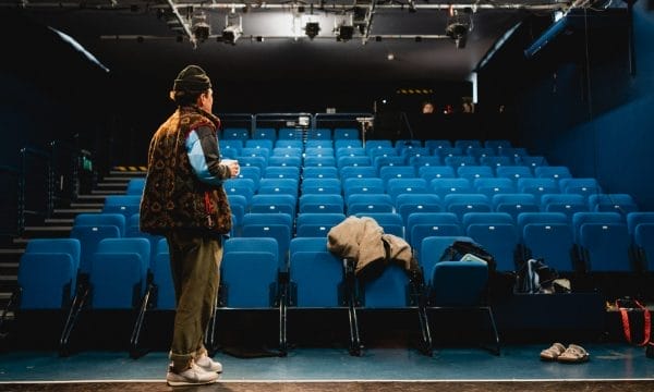 A person stands on stage facing empty blue theater seats, with some coats and a bag left on the chairs and floor.