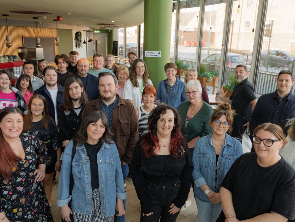 Team of approximately 35-40 people gathered for a group photo in a bright, modern office space featuring green pillars, wooden slat walls, and floor-to-ceiling windows overlooking the street