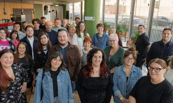 Team of approximately 35-40 people gathered for a group photo in a bright, modern office space featuring green pillars, wooden slat walls, and floor-to-ceiling windows overlooking the street