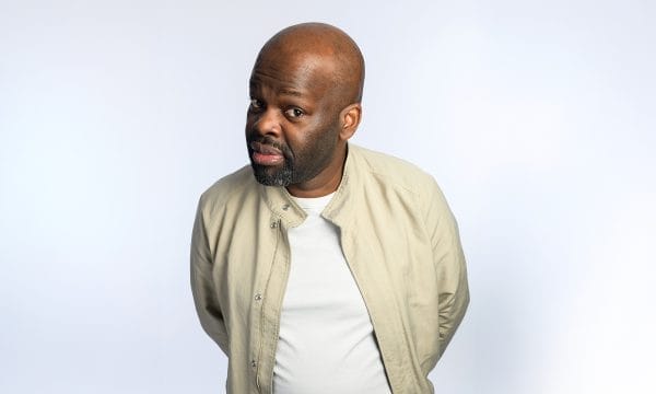 Daliso is a black male with a bald head and shortly shaven black beard. He is wearing a white tshirt and cream coat.