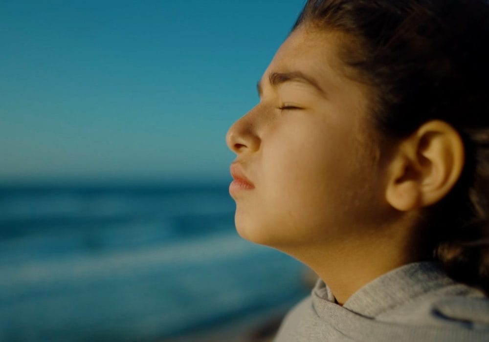 A captivating image displays a young girl, serenely standing on a sunlit beach, her eyes closed. Gray sweatshirt, tousled bun, and the oceans gentle embrace create a peaceful, contemplative scene.