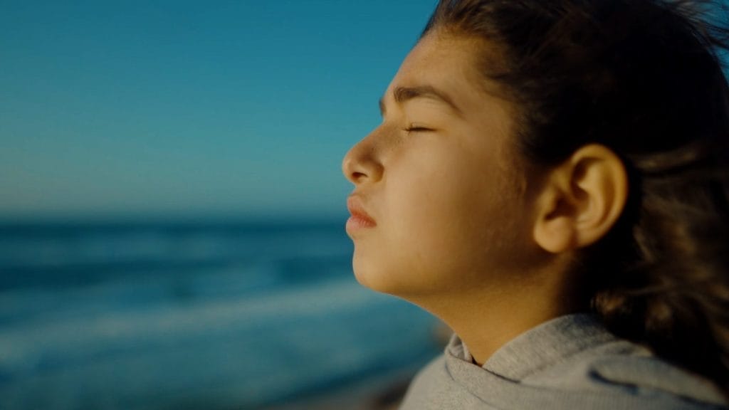 A captivating image displays a young girl, serenely standing on a sunlit beach, her eyes closed. Gray sweatshirt, tousled bun, and the oceans gentle embrace create a peaceful, contemplative scene.