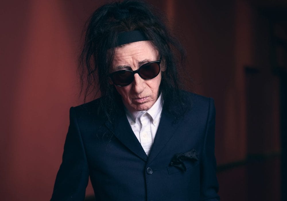 John is a person with long dark hair wearing dark sunglasses and a navy blue suit jacket with white shirt underneath. They have a serious expression and are photographed against a dark reddish-brown background with dramatic lighting.