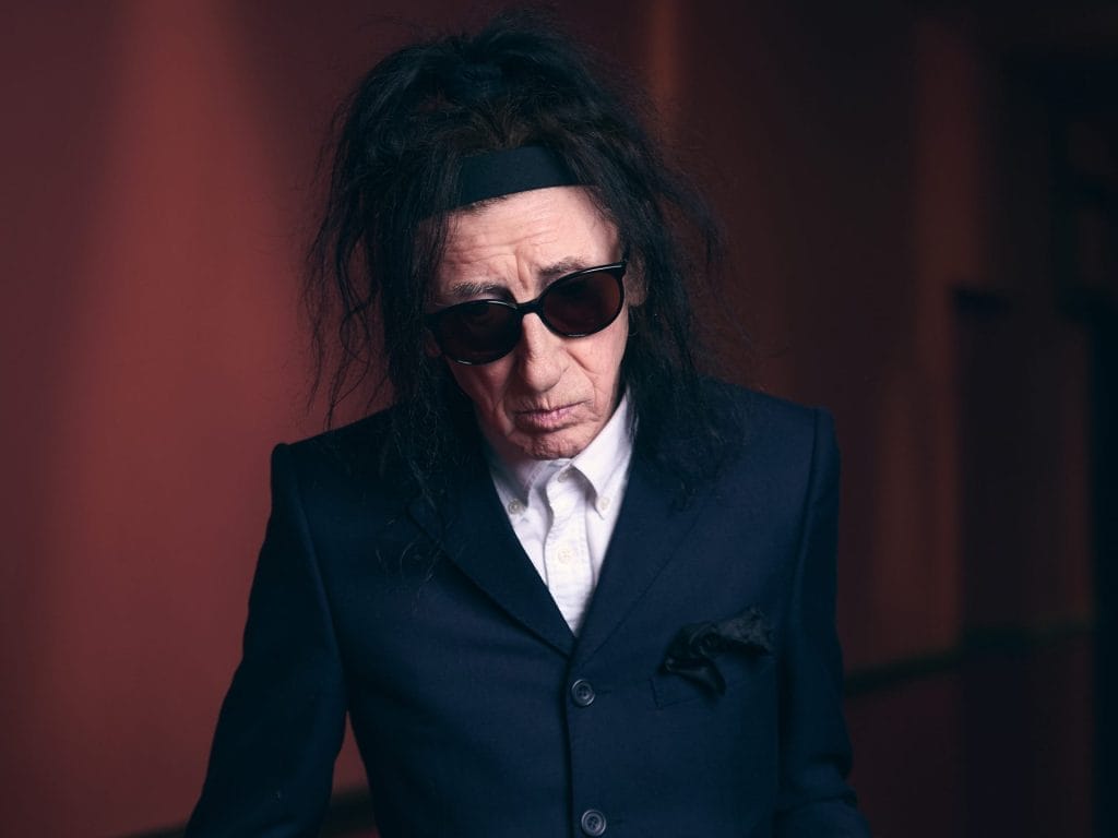 John is a person with long dark hair wearing dark sunglasses and a navy blue suit jacket with white shirt underneath. They have a serious expression and are photographed against a dark reddish-brown background with dramatic lighting.