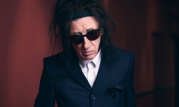 John is a person with long dark hair wearing dark sunglasses and a navy blue suit jacket with white shirt underneath. They have a serious expression and are photographed against a dark reddish-brown background with dramatic lighting.