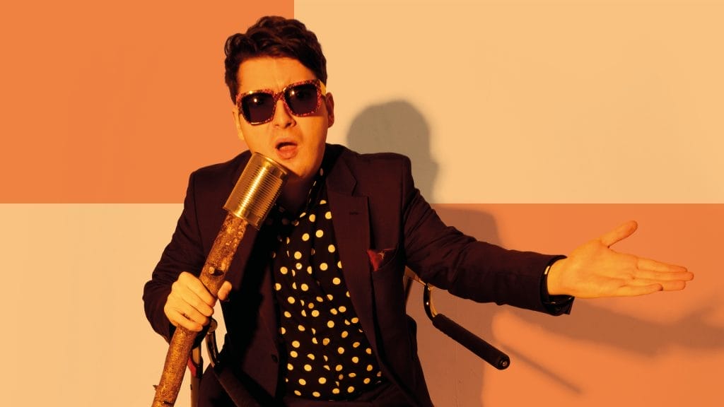 A captivating image displays a dapper man in a black suit with polka dots, sunglasses, and a microphone. His dynamic pose against an orange-white gradient background captivates, highlighting a skilled performer in peak expression.