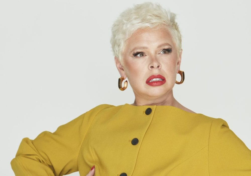 Laura is a person wearing a mustard jacket. She has short white-blonde hair wearing big gold hoops. She has her hands on her hips looking away from the camera.