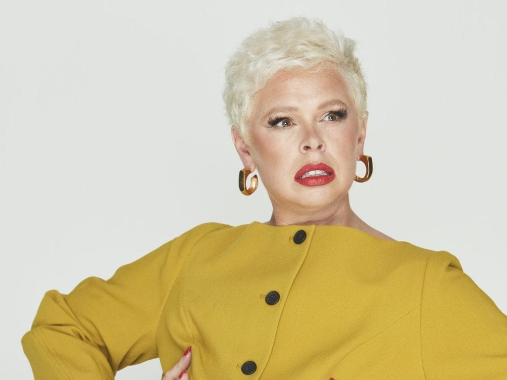 Laura is a person wearing a mustard jacket. She has short white-blonde hair wearing big gold hoops. She has her hands on her hips looking away from the camera.