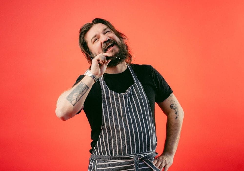 Matt holds a plastic spoon used for cooking next to his head and talks into it like a phone while wearing an apron.