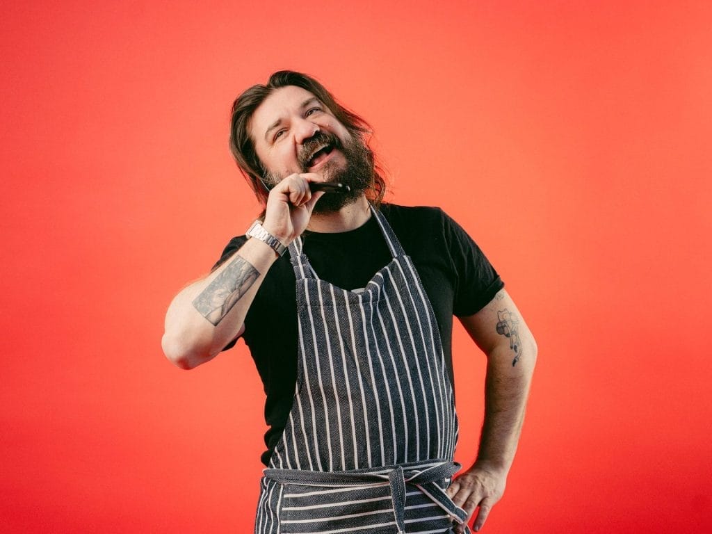 Matt holds a plastic spoon used for cooking next to his head and talks into it like a phone while wearing an apron.