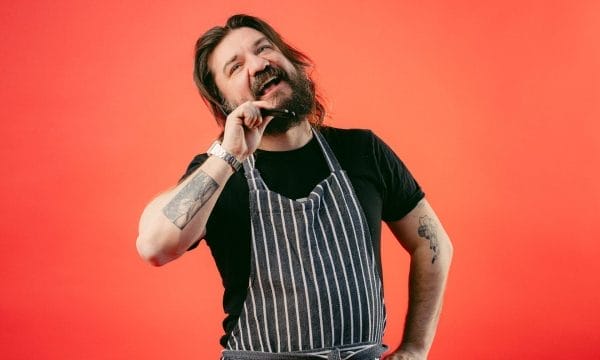 Matt holds a plastic spoon used for cooking next to his head and talks into it like a phone while wearing an apron.