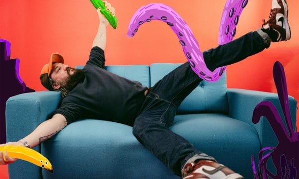 A person lying on a light blue sofa against a vibrant orange-red background, appearing to be grabbed by large purple tentacles with suction cups. They're holding a green pickle-shaped object and wearing dark clothing with trainers. Yellow and purple tentacle-like shapes frame the scene, creating a surreal, playful composition that blends reality with fantastical elements.