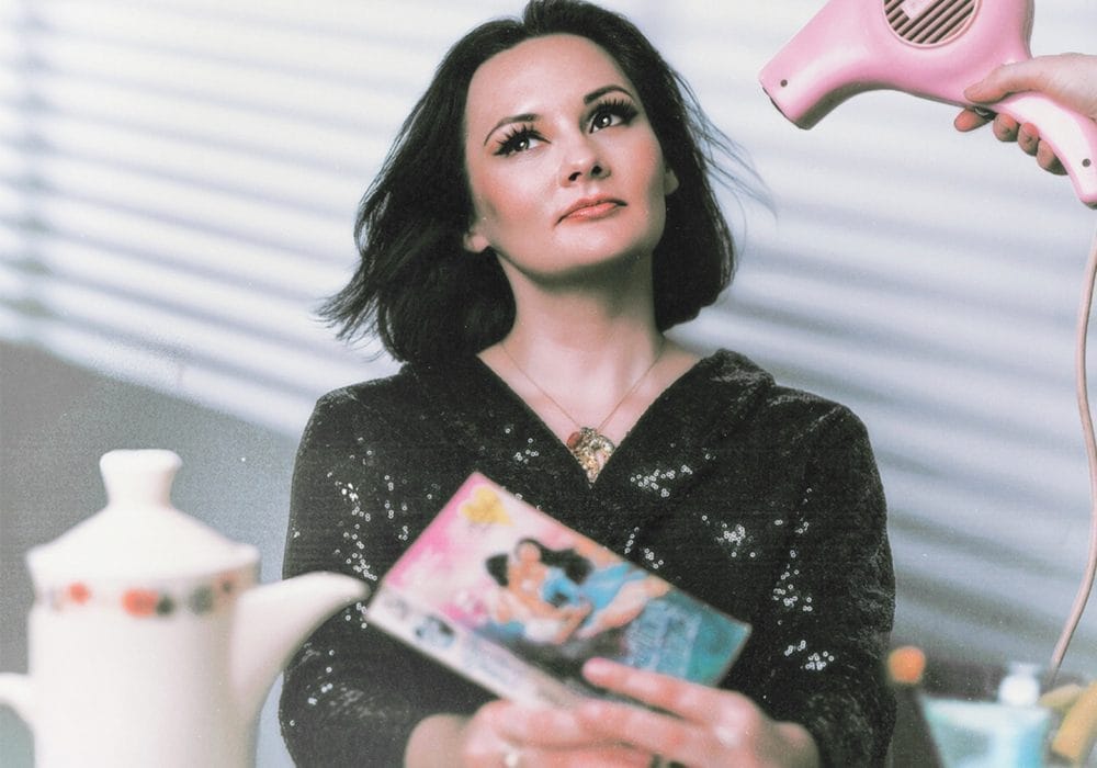 A woman with dark hair in a black dress sits at a table holding a vintage magazine. She faces upward as a pink hairdryer is aimed at her. On the table are a teapot, teacup, and retro kitchen items.