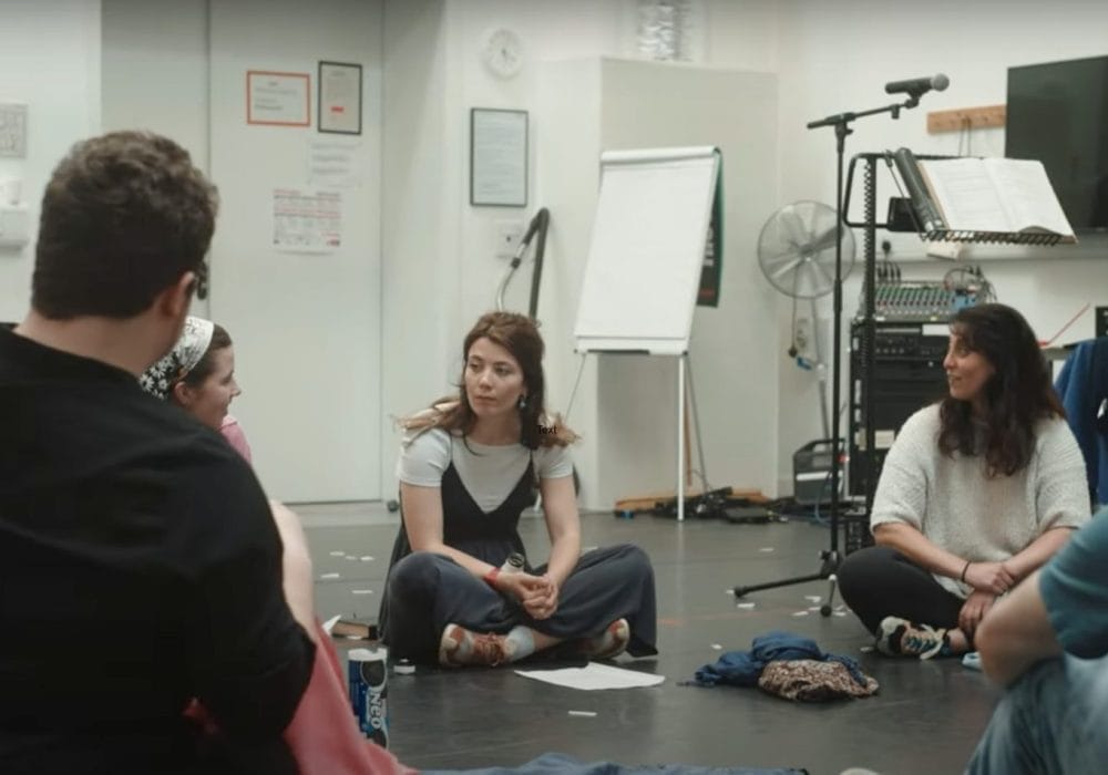 A group of people sit in a circle on the floor of a rehearsal room, engaged in conversation, with musical equipment and papers scattered around.