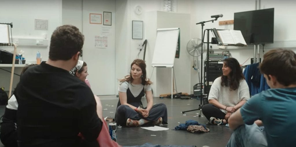 A group of people sit in a circle on the floor of a rehearsal room, engaged in conversation, with musical equipment and papers scattered around.