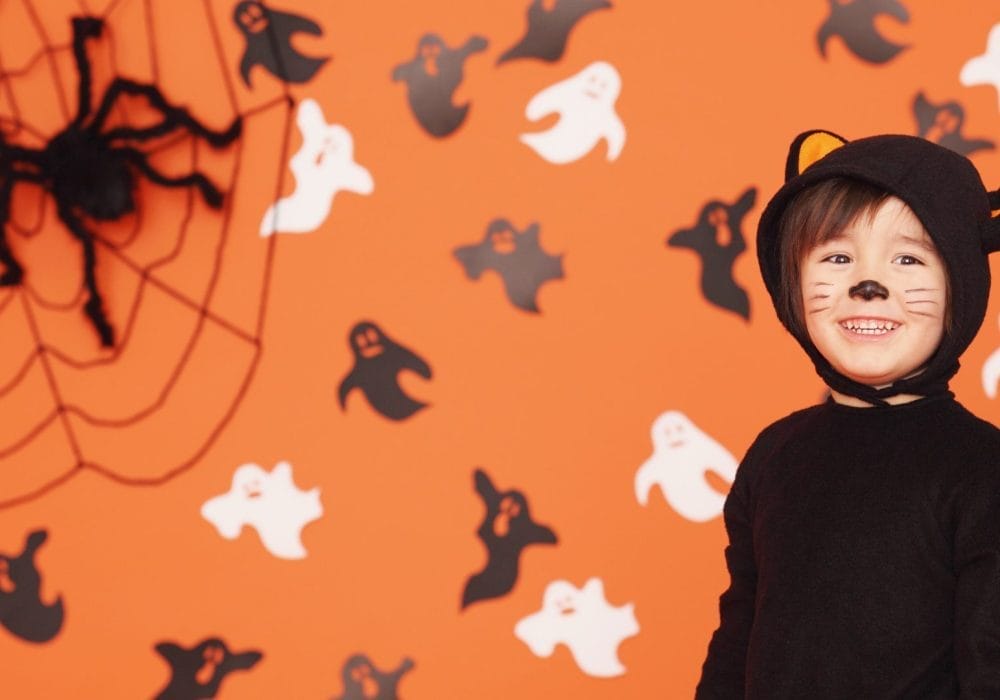 Child in a black cat costume with face paint smiles in front of an orange wall decorated with ghosts and a large spider web.