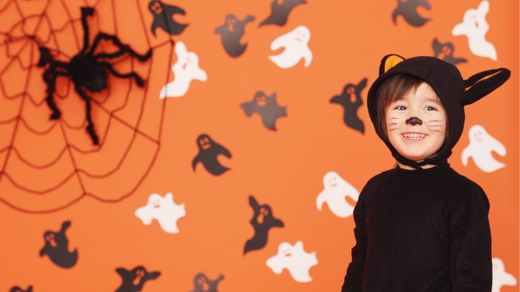 Child in a black cat costume with face paint smiles in front of an orange wall decorated with ghosts and a large spider web.