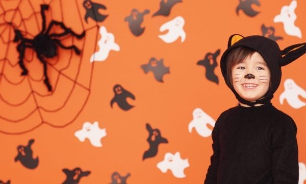 Child in a black cat costume with face paint smiles in front of an orange wall decorated with ghosts and a large spider web.