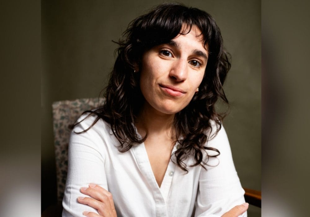 Portrait of a woman with long, dark, wavy hair and a white button-down shirt, arms crossed. She is seated in a patterned chair against a green-toned background, looking directly at the camera with a slight smile.