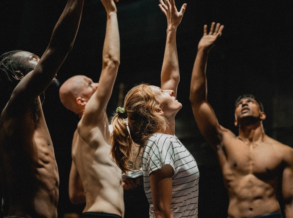 A group of people with their arms raised upward, reaching toward light from above. The image shows several individuals including a woman in a striped shirt in the centre, with men around her, some shirtless. They are photographed from a low angle against a dark background with dramatic lighting creating strong contrast. Their faces are tilted upward and their arms stretched high, creating a sense of collective aspiration or yearning.