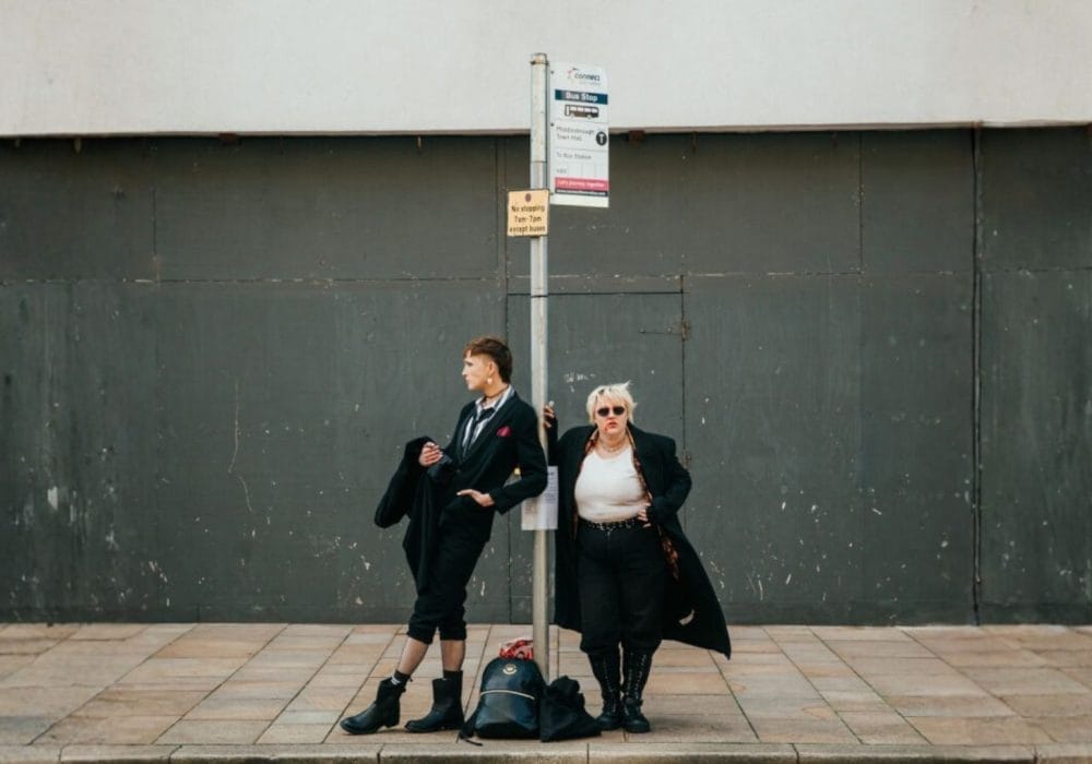 Two people stand at a bus stop in front of a gray wall; one is adjusting their coat while the other looks off to the side with hands on hips.