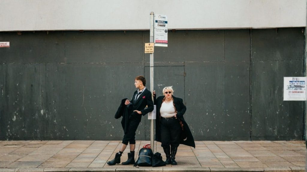 Two people stand at a bus stop in front of a gray wall; one is adjusting their coat while the other looks off to the side with hands on hips.