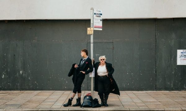 Two people stand at a bus stop in front of a gray wall; one is adjusting their coat while the other looks off to the side with hands on hips.