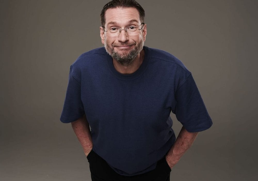 A mature man with brown hair, short beard and glasses wearing a dark blue t-shirt leaning towards the camera with a smug look on his face
