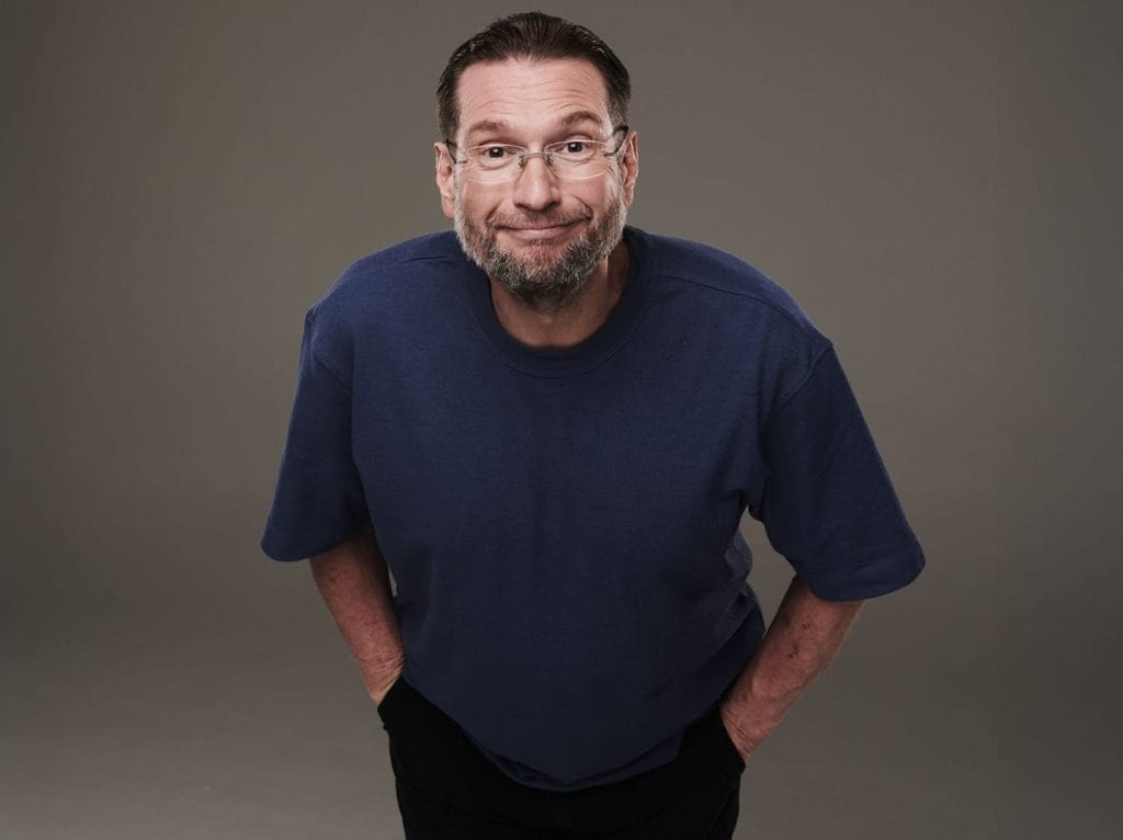 A mature man with brown hair, short beard and glasses wearing a dark blue t-shirt leaning towards the camera with a smug look on his face