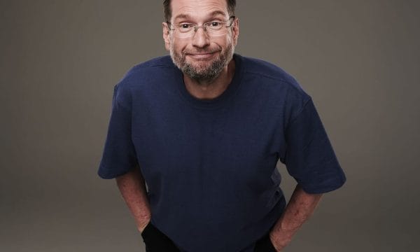 A mature man with brown hair, short beard and glasses wearing a dark blue t-shirt leaning towards the camera with a smug look on his face