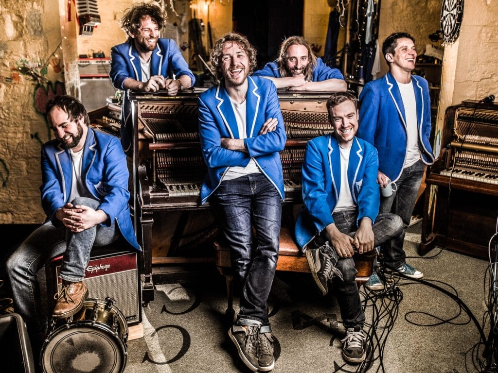 A group of 6 musicians wearing blue jackets resting casually on a piano and amps