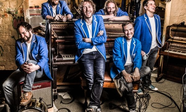 A group of 6 musicians wearing blue jackets resting casually on a piano and amps