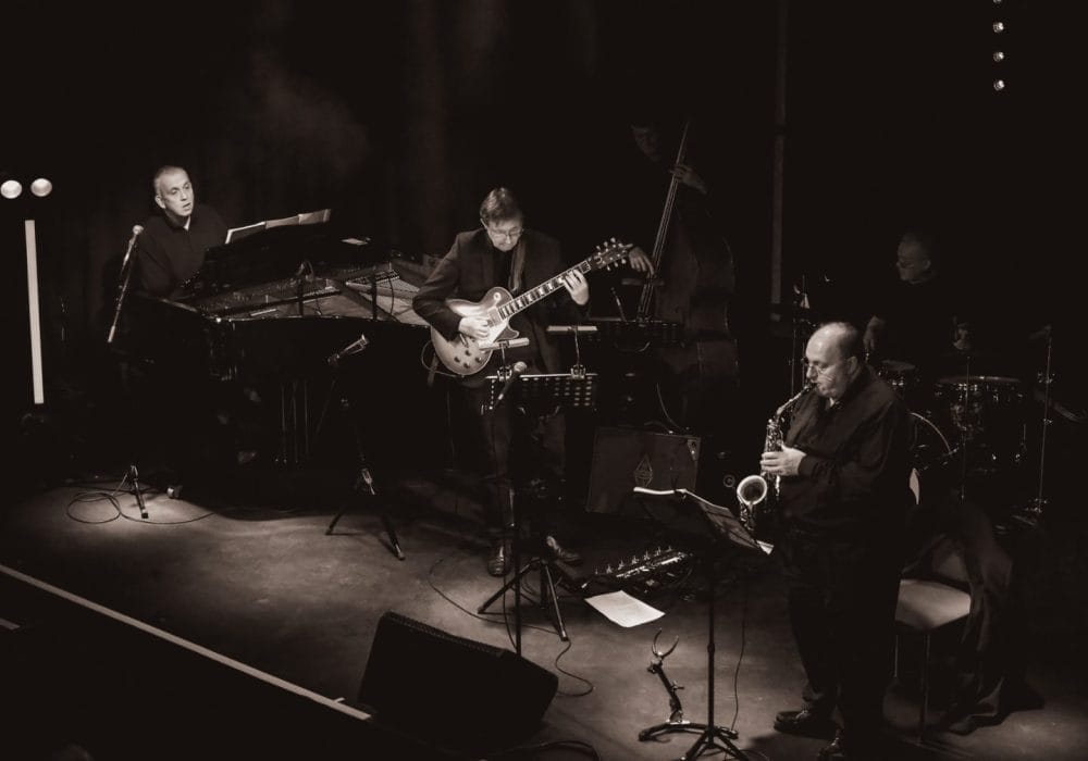 Black and white image of musicians playing together on stage, showcasing their instruments and dynamic performance.