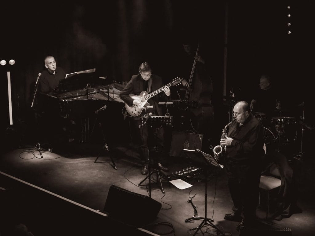 Black and white image of musicians playing together on stage, showcasing their instruments and dynamic performance.